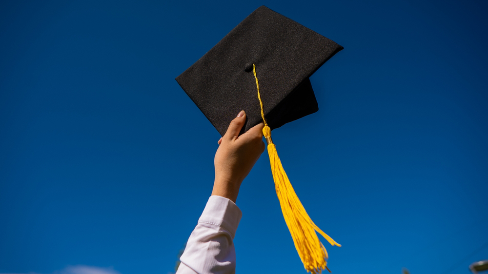 Hand holding a black graduation cap with a yellow tassel against a blue sky.